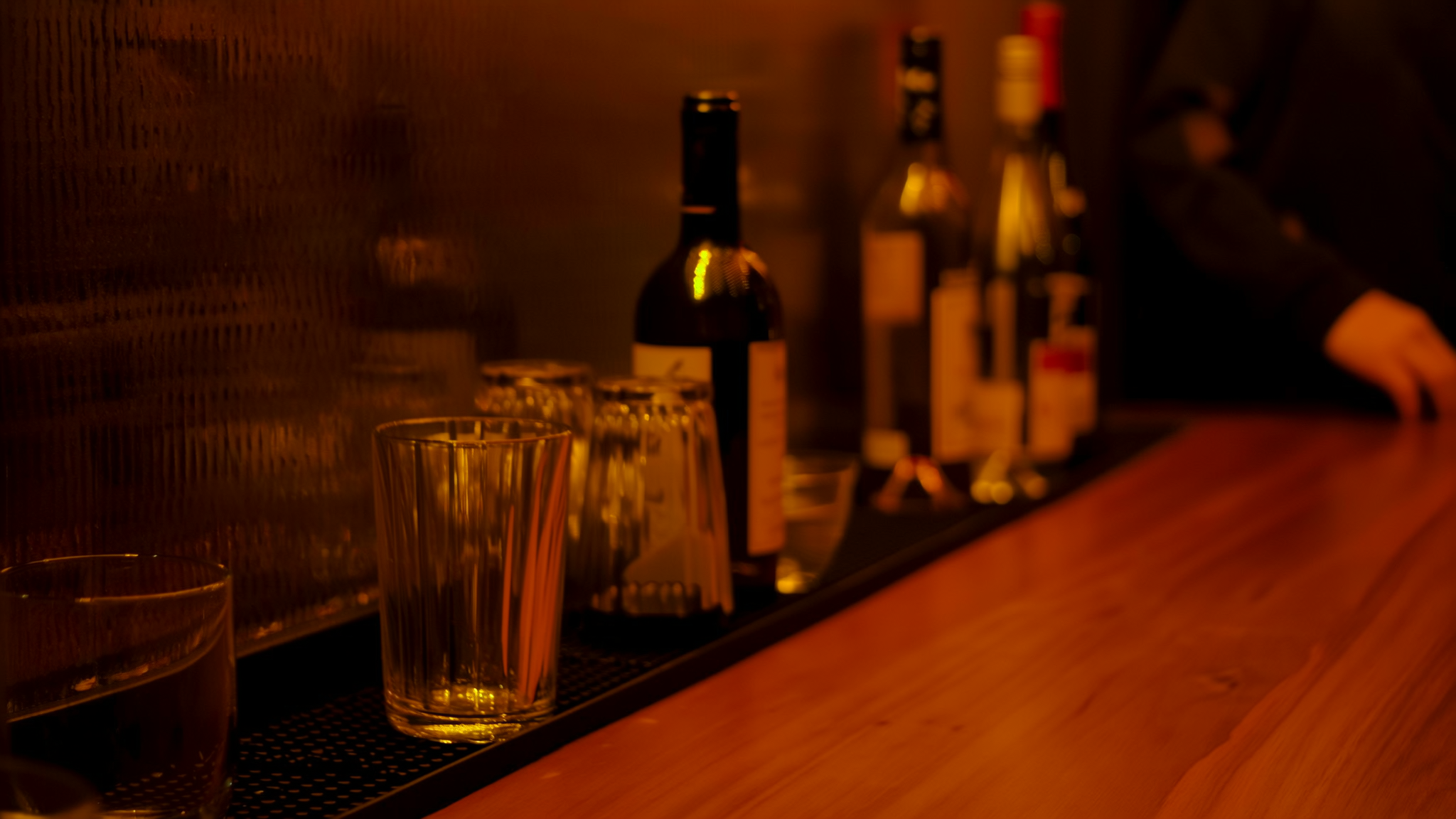 Flessen en glazen op een houten bar, warm verlicht in de avond.