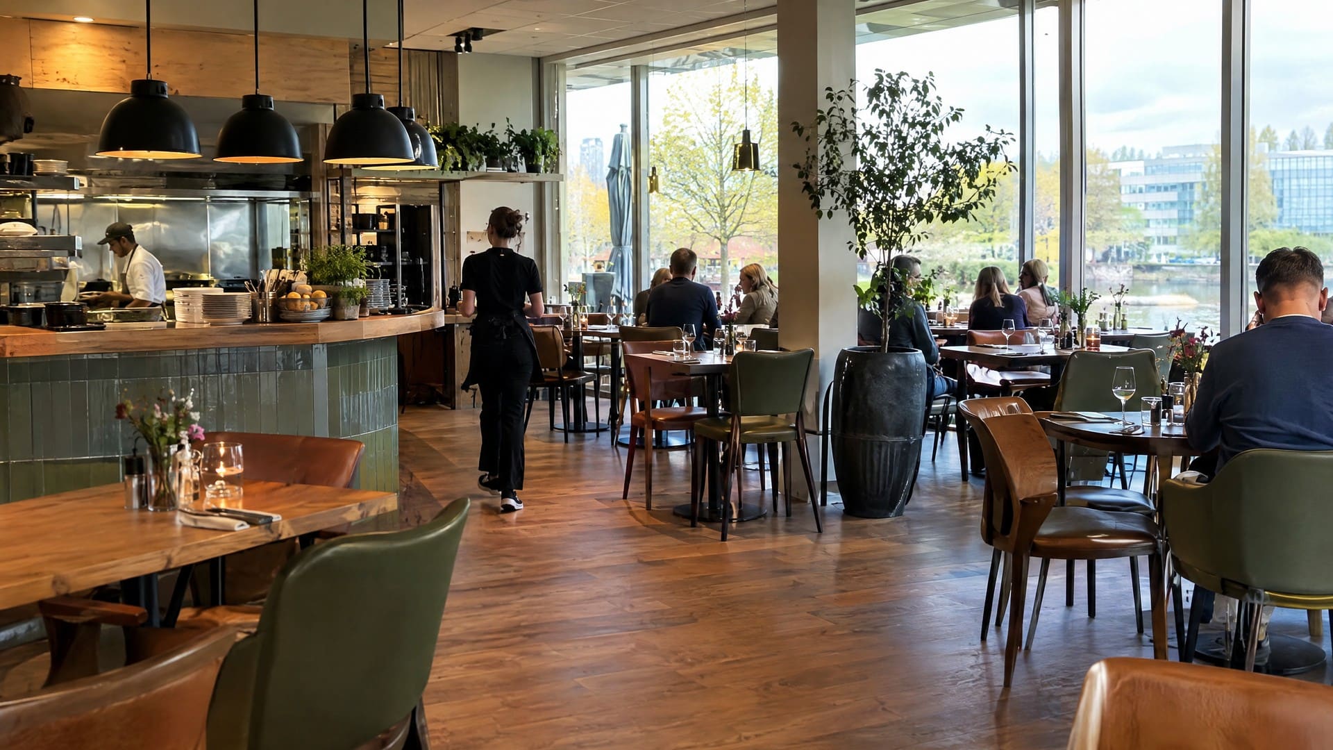 Het verlichte interieur van een bar bistro: een open keuken achter een groen betegelde toonbank, houten tafels met leren stoelen en hoge ramen die uitkijken op het water.